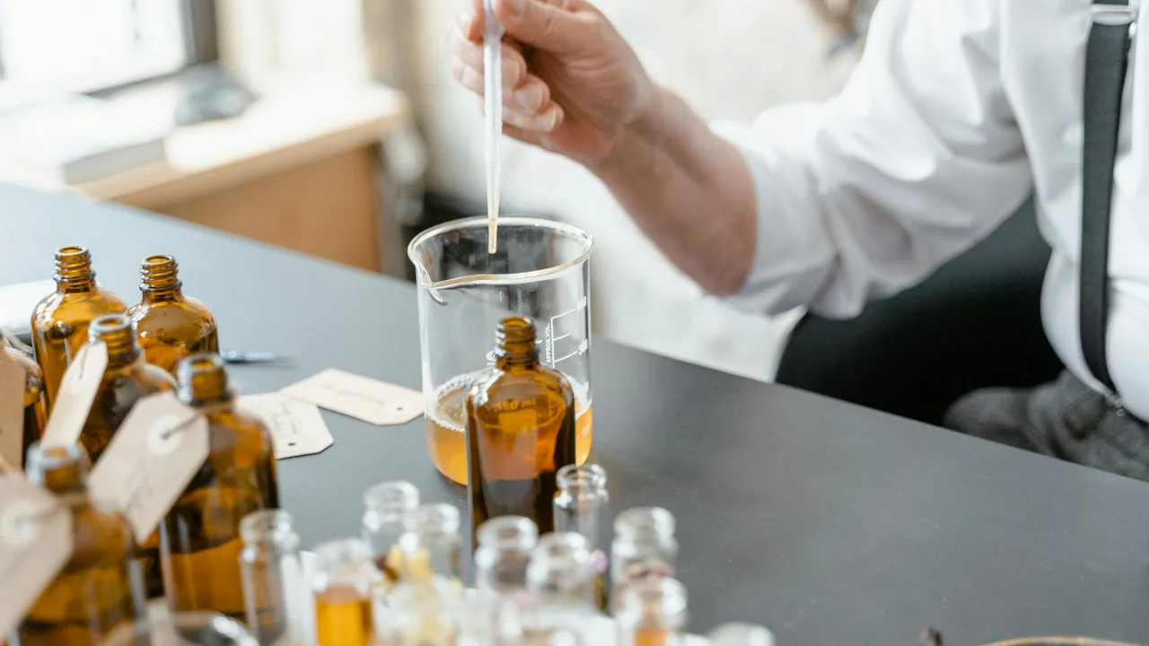 A researcher or technician uses a dropper over a glass beaker on a laboratory bench, with several amber essential oil bottles nearby.