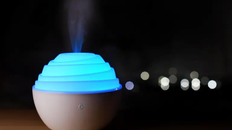 A white, spherical essential oil diffuser with a glowing blue top emitting mist at night, against a dark background with blurred city lights.