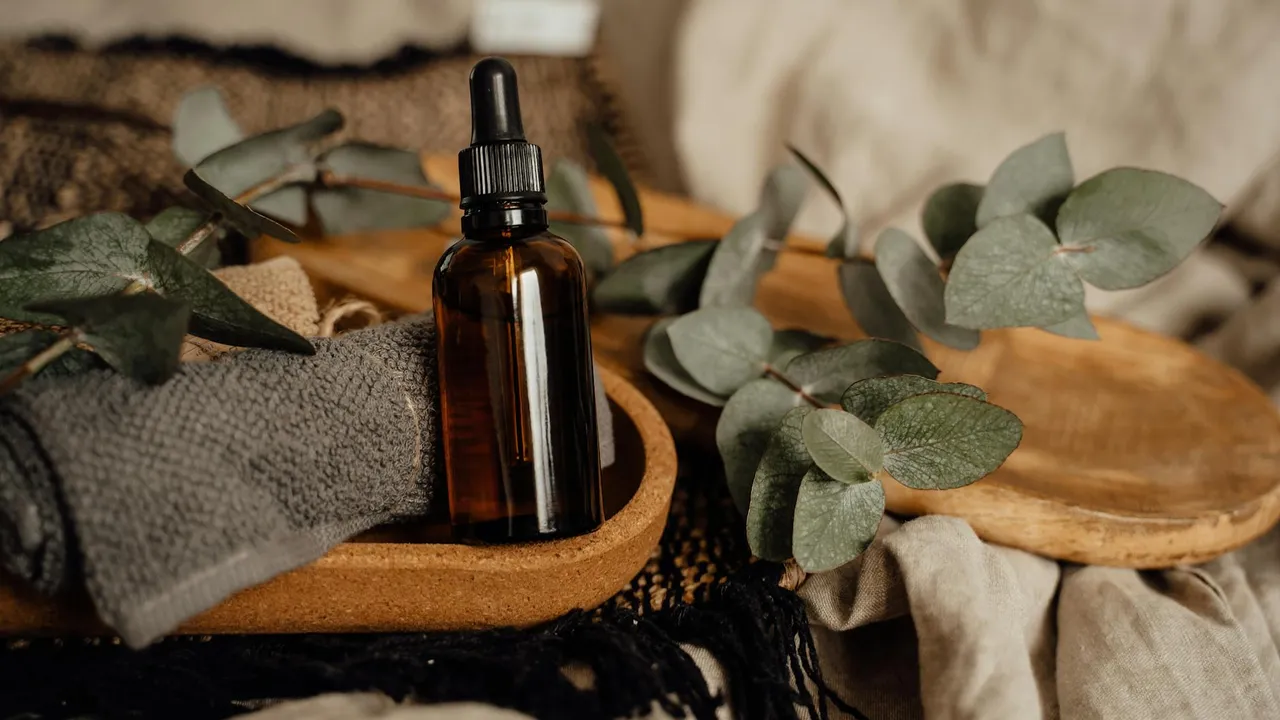 A small amber glass dropper bottle placed on a wooden surface, surrounded by eucalyptus leaves, a gray cloth, and wooden massage slippers, suggesting a spa-like oil setup.