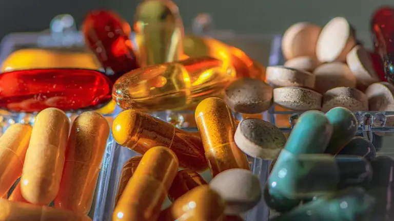 Close-up of assorted supplements: yellow and orange fish oil capsules, green and blue tablets, and brown pills scattered on a cool-toned surface.
