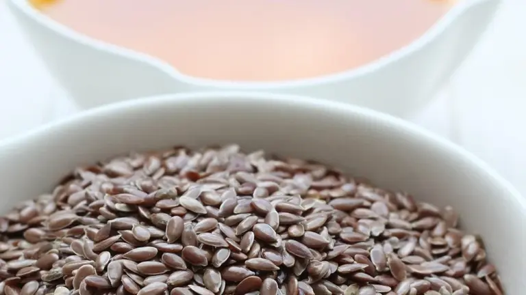 Close-up of a white bowl filled with flax seeds with a blurred orange sauce in the background.