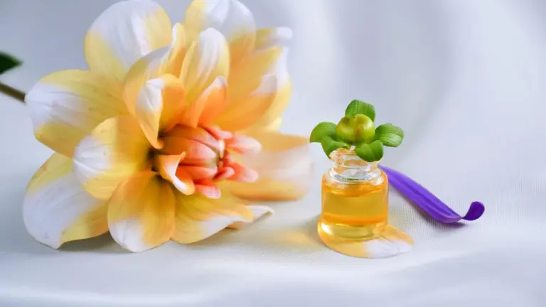 Close-up of a pale-yellow flower and a small bottle of golden body oil with a green leaf on top, resting on a white satin surface with a purple ribbon in the background.