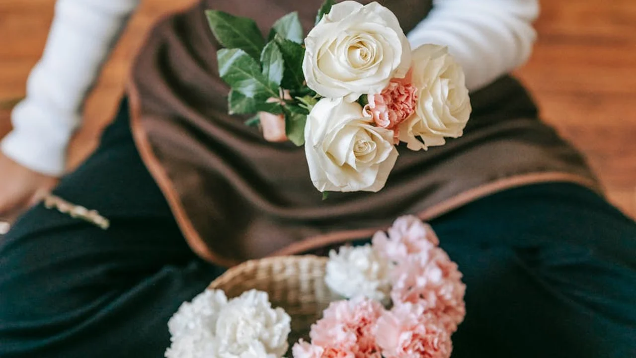 A person wearing an apron holds a bouquet of white and pale pink roses, with a basket of flowers nearby, suggesting natural ingredients for DIY perfumes and colognes.