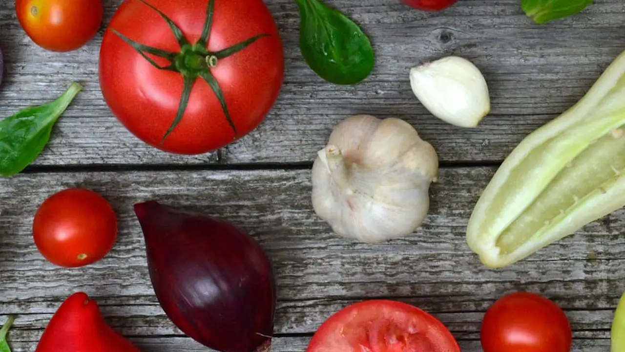 Assorted fresh vegetables on a rustic wooden surface, including tomatoes, garlic, red onion, basil, and a pale green stalk.