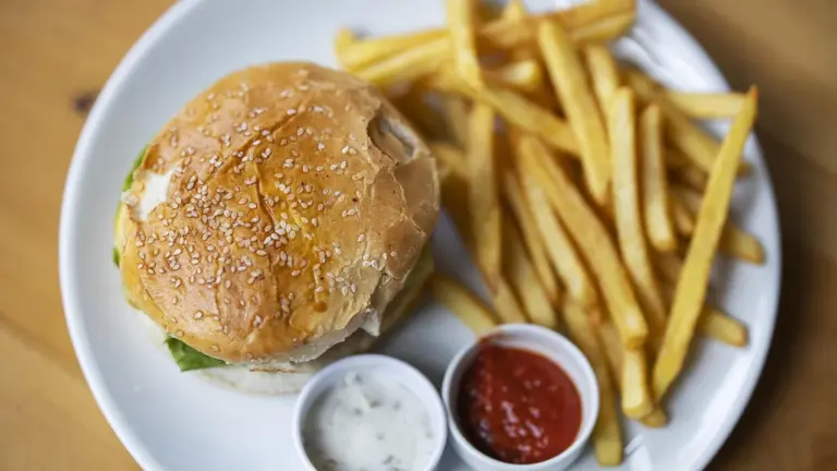 Burger with sesame seed bun and a portion of french fries on a white plate with two dipping sauces.