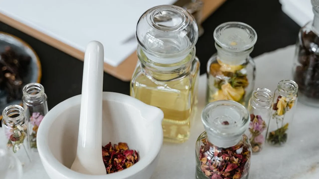 Home apothecary setup with a white mortar and pestle, small glass bottles of dried botanicals, and jars of infused oil on a dark work surface.