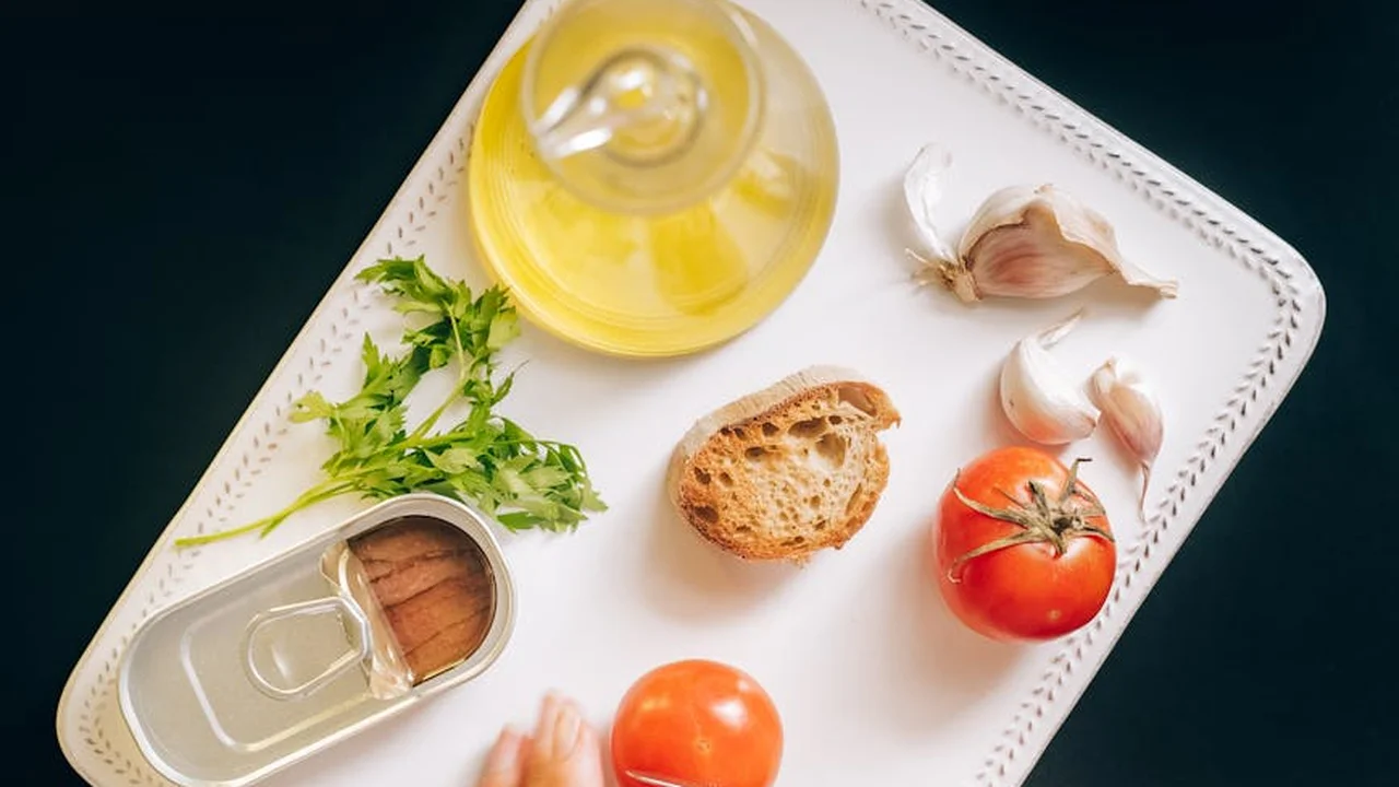 A white tray holds garlic cloves, a bottle of olive oil, parsley, a tomato, bread, and other ingredients laid out for infusing garlic into olive oil.