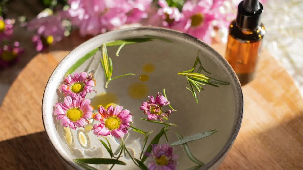 A metal bowl of water with floating pink daisy-like flowers and rosemary sprigs; an amber essential oil bottle sits on a wooden surface in the background.