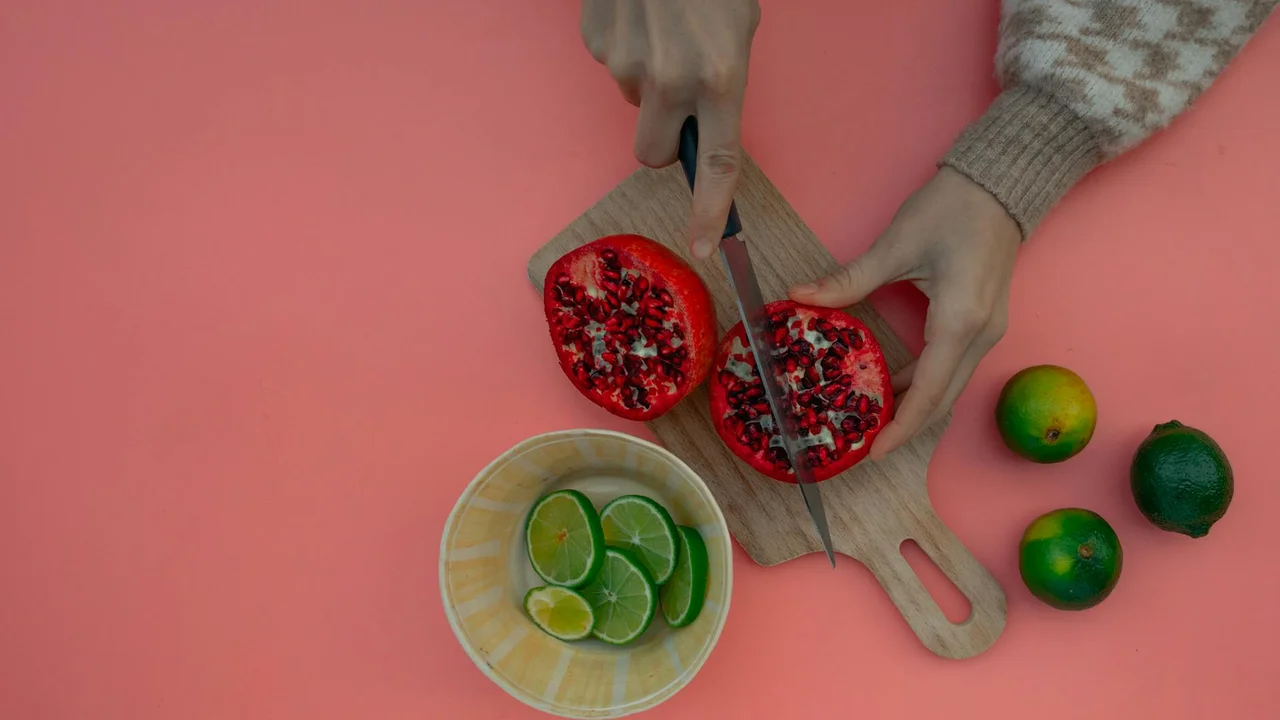 Hands with a knife cutting red pomegranate halves on a wooden cutting board, with lime wedges in a small bowl and whole limes on a pink background