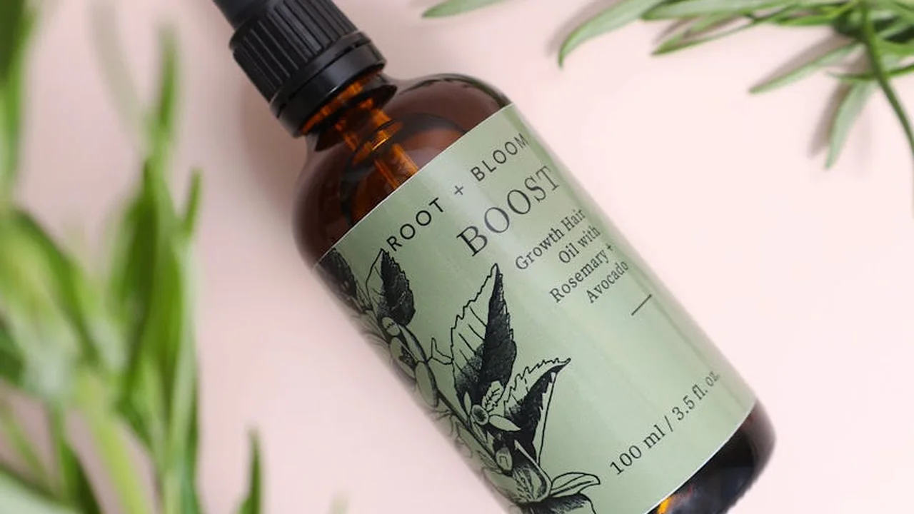 Bottle of hair oil with a botanical label on a soft pink backdrop, surrounded by green leaves.