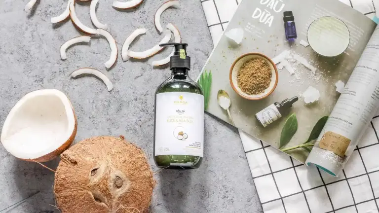 Flat lay of hair care ingredients: a bottle of olive oil with a pump, coconut pieces, coconut shavings, and a beauty magazine on a textured gray surface.