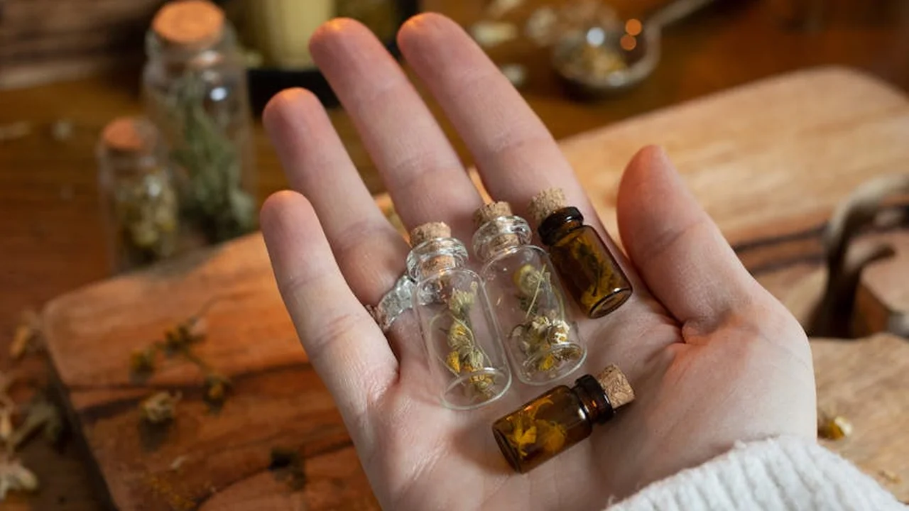 Close-up of a hand holding several small glass vials of essential oils, with herbs and a wooden surface in the background