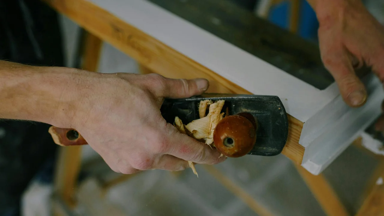 Close-up of hands using a hand plane to smooth a wooden board in a workshop.