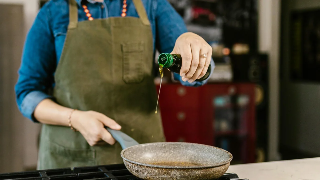 Person in a green apron pours oil into a skillet, with a kitchen backdrop.