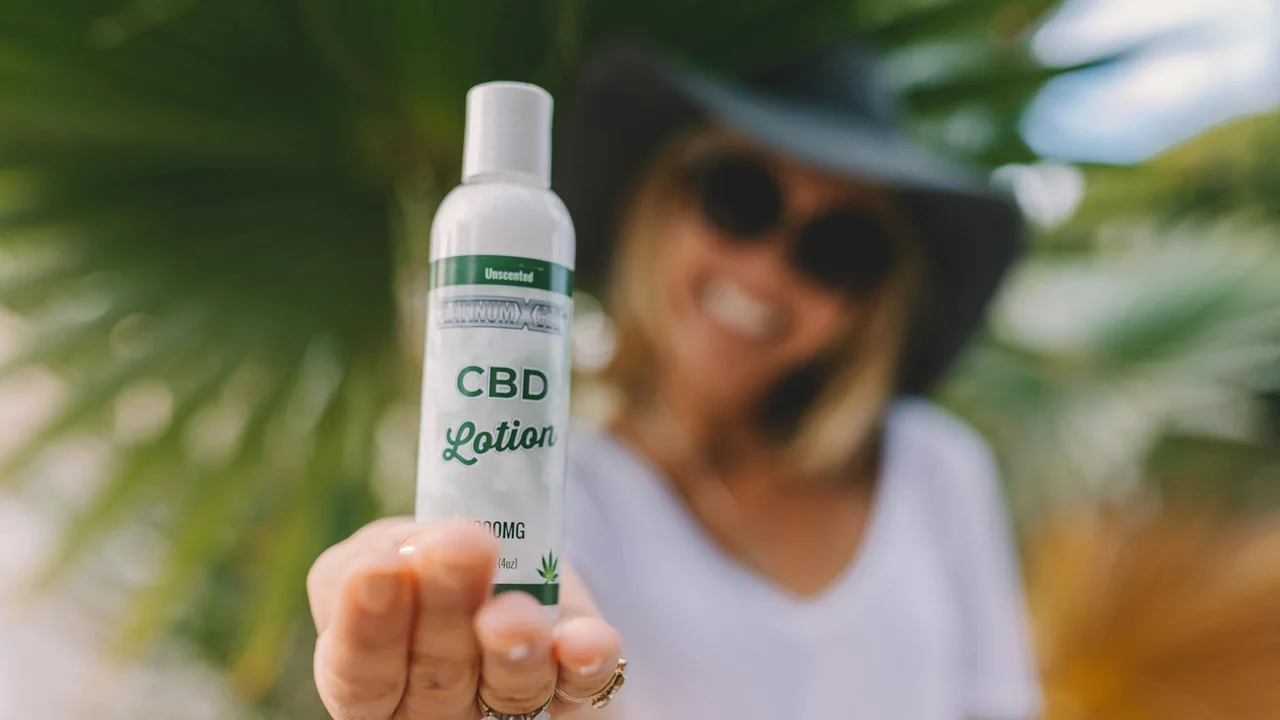 A person wearing a sunhat and sunglasses holds a bottle labeled CBD Lotion in the foreground, with a sunny outdoor setting in the background.