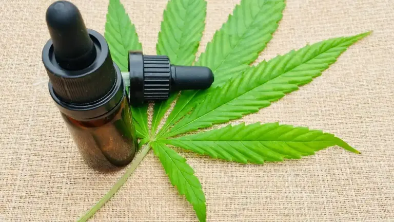 Dropper bottle of hemp oil placed beside a cannabis leaf on a beige fabric background.