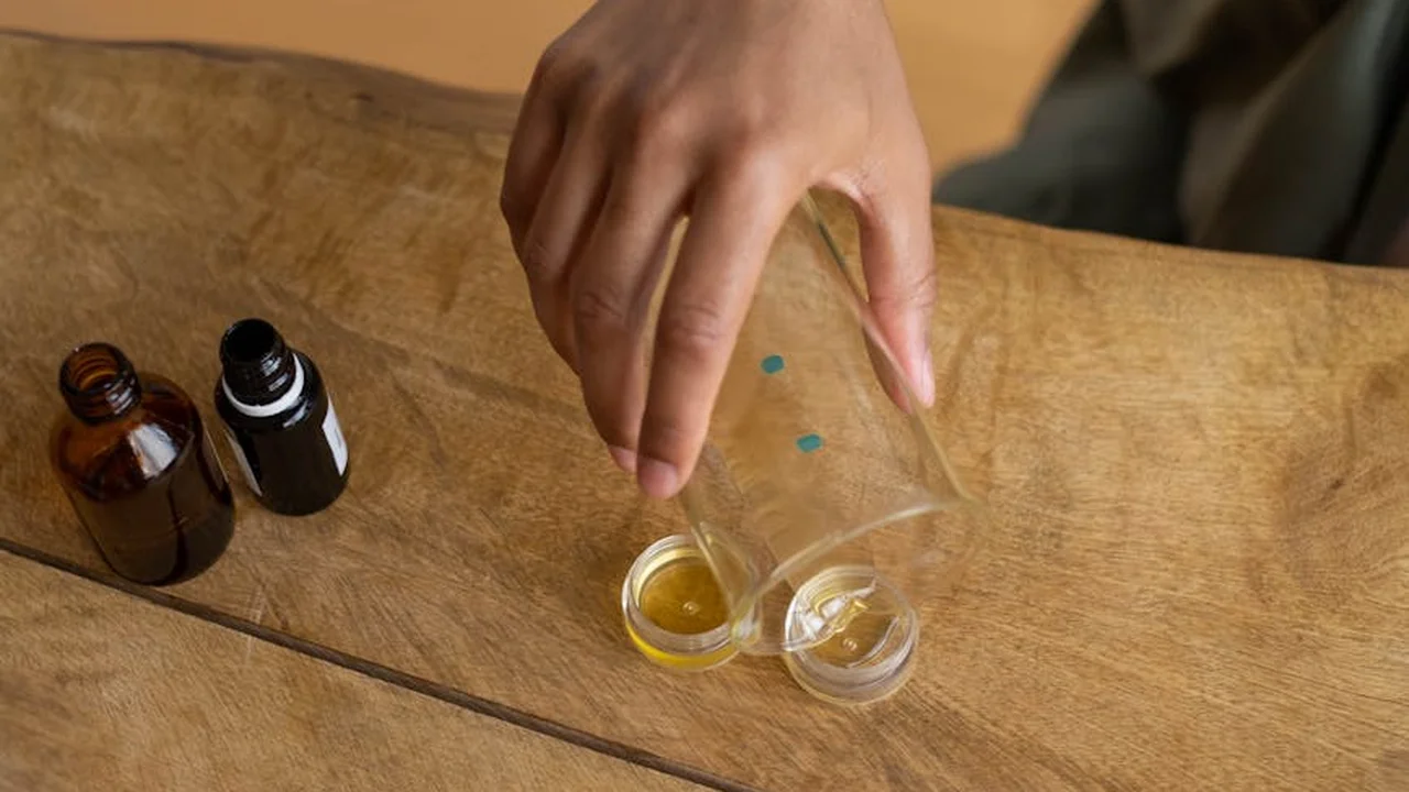 A person’s hand pouring oil into a small measuring cup on a wooden table, with two brown glass bottles and other containers nearby, illustrating preparation for making herbal infused oils at home.