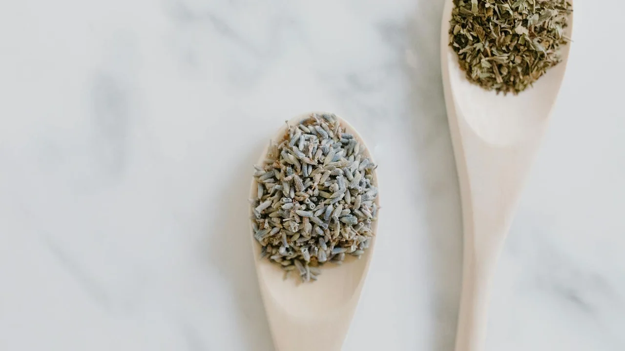 Two wooden spoons filled with dried herbs on a marble surface, ready for herbal oil infusion.