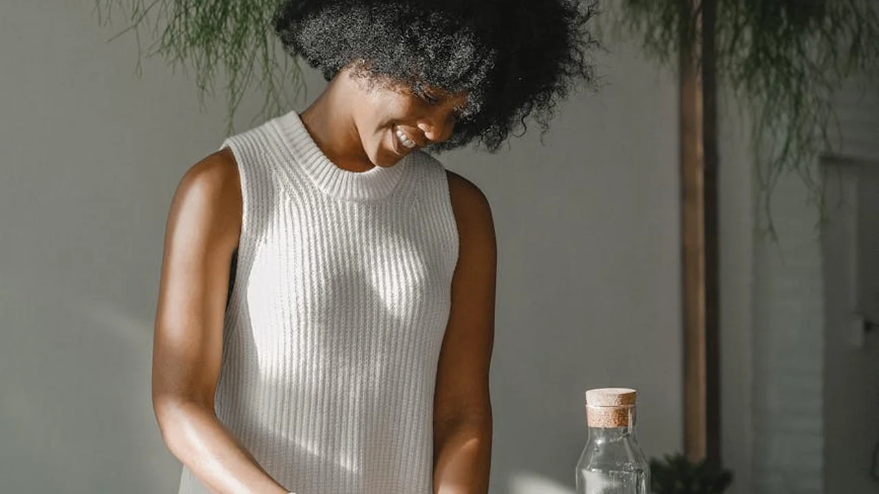 A smiling woman in a sleeveless white knit top stands indoors beside lush greenery, illustrating the human side of botanical oil extraction.