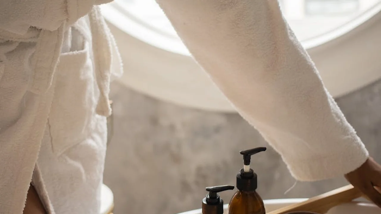 Close-up of a person in a robe reaching for a pump bottle on a wooden surface in a bright, spa-like setting.