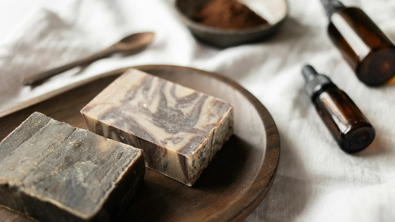 Two handmade soaps on a round wooden plate with small amber essential oil bottles in the background, set on soft light fabric.
