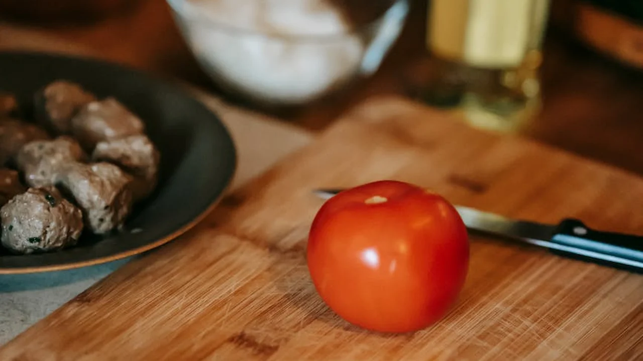 Red tomato on a wooden cutting board with a knife nearby; a skillet with meat bites blurred to the left and a bottle of oil in the background.
