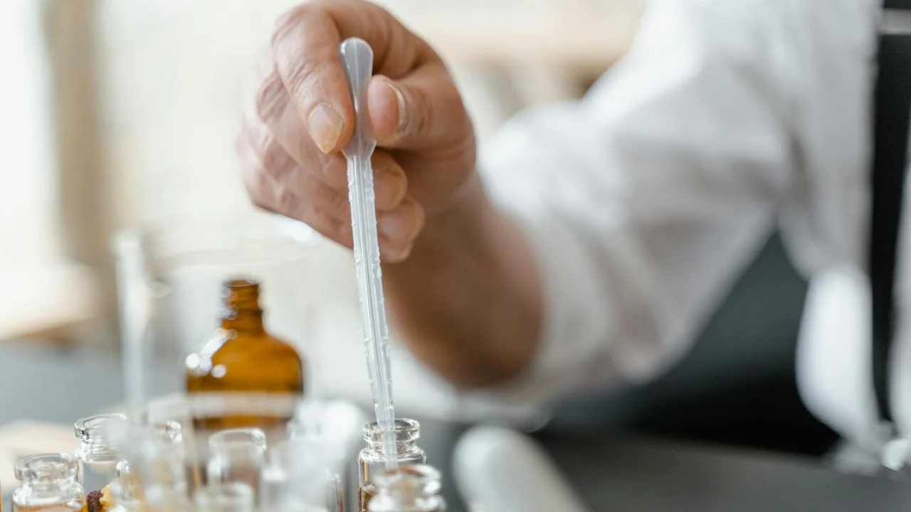 Close-up of a hand holding a dropper over several small amber bottles of botanical oil in a lab-like setting.