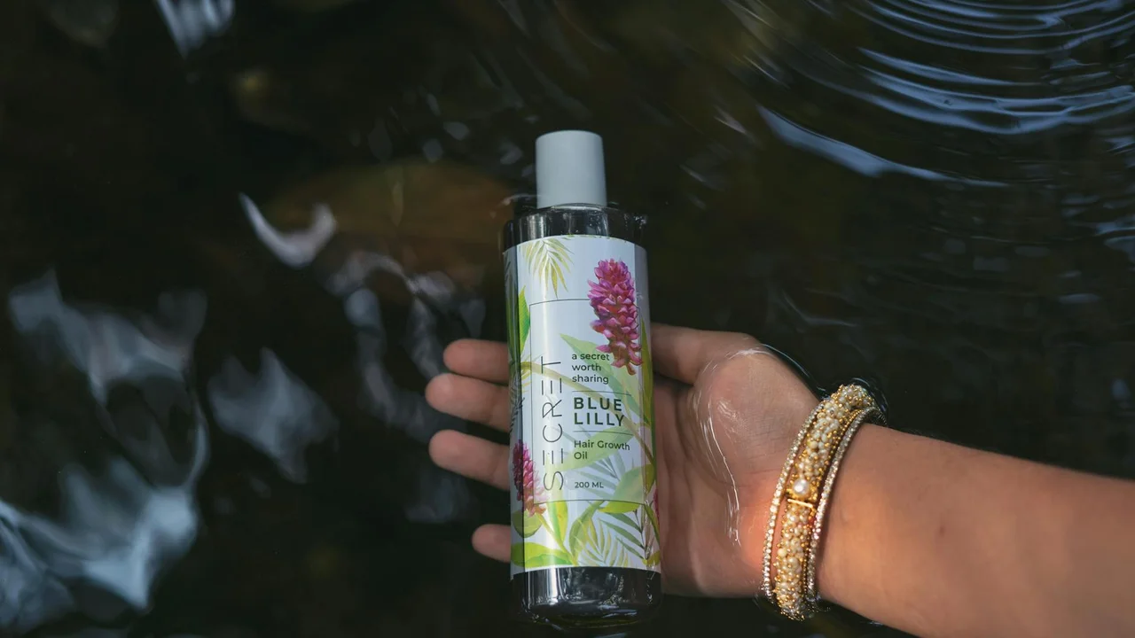 A hand with gold bracelets holding a bottle of hair oil with a botanical label, against a blurred water-reflection background.