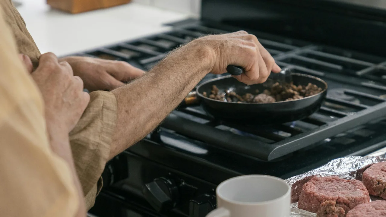 Hands using tongs to fry food in a skillet on a gas stove; raw burger patties sit nearby on the counter.