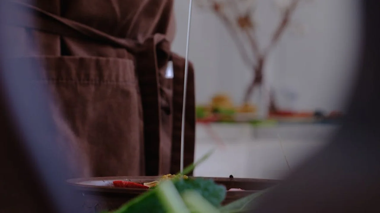A person wearing a brown apron stands in a home kitchen, with a plate and knife visible in the foreground and a blurred kitchen background.