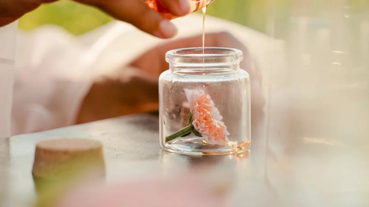 Hand holding a dropper over a small glass jar, releasing oil onto plant material inside, with a soft, blurred background.