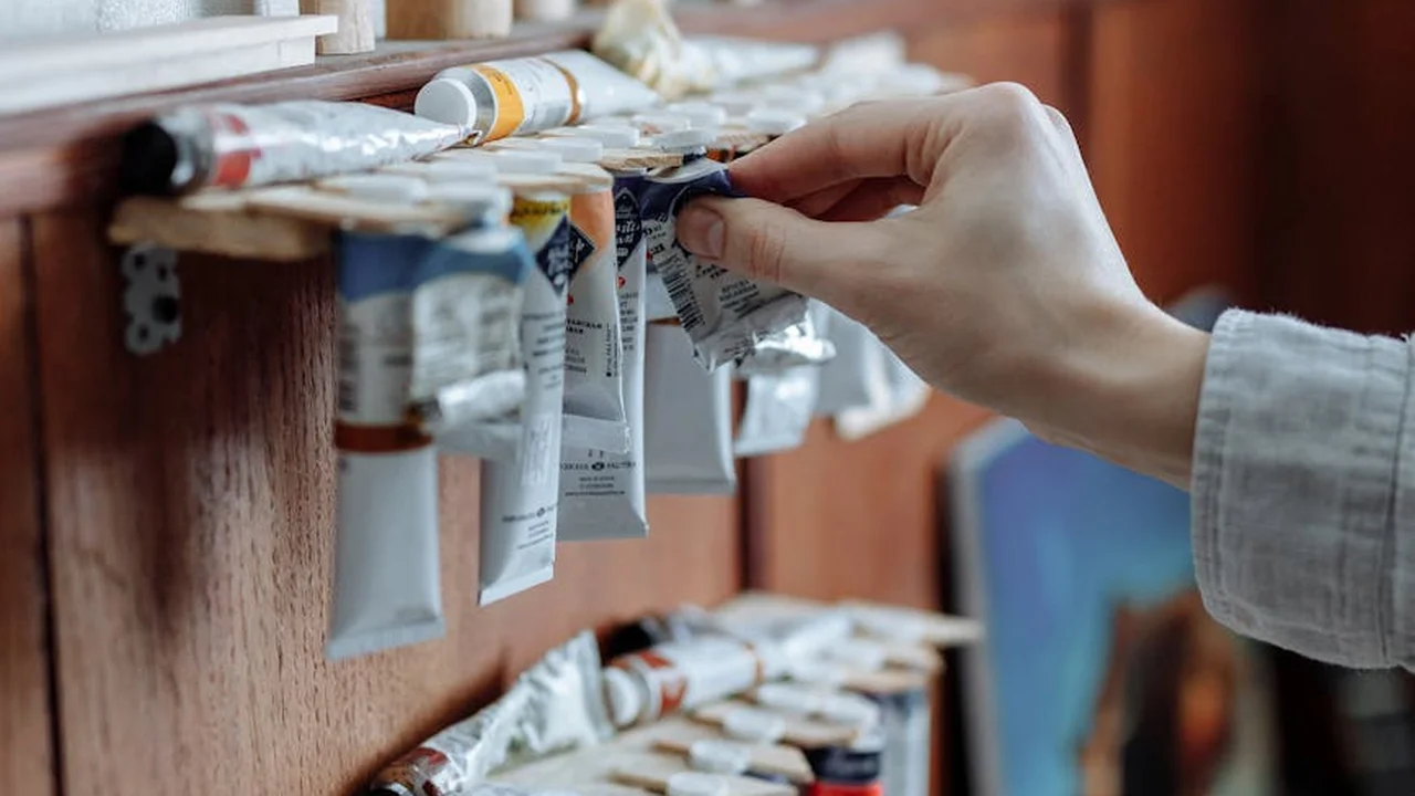 Close-up of a hand selecting a tube of oil paint from a rack of paint tubes on a wooden shelf.