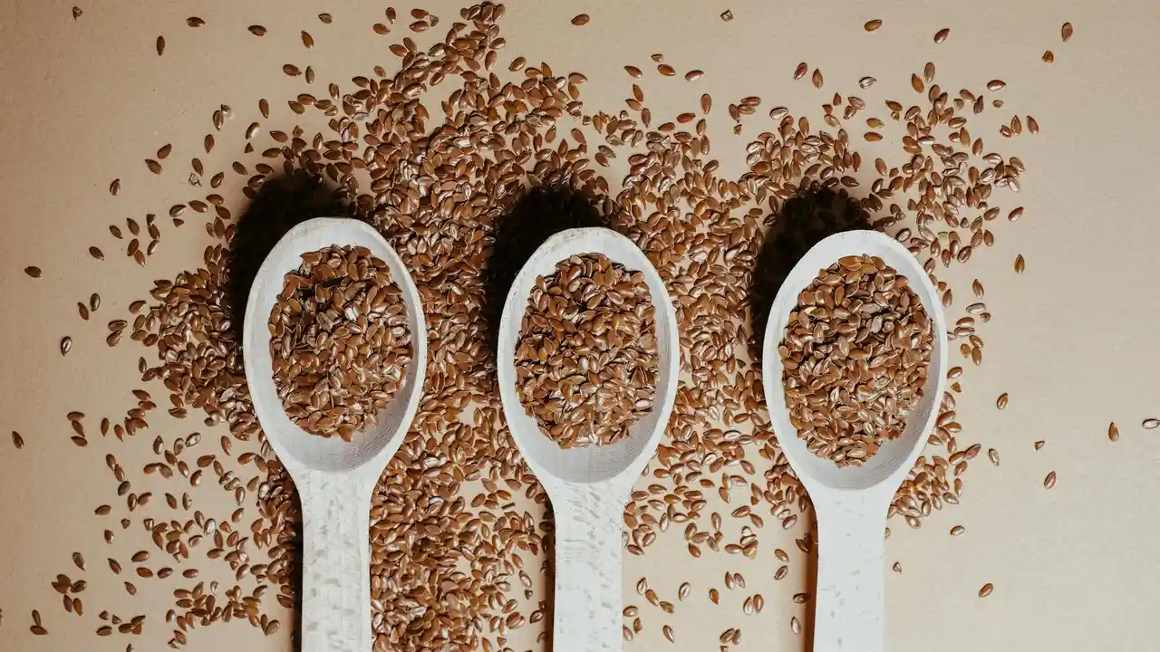 Three white spoons filled with flax seeds, with loose seeds scattered on a light background, illustrating flax (linseed) used for oil production in historical contexts.