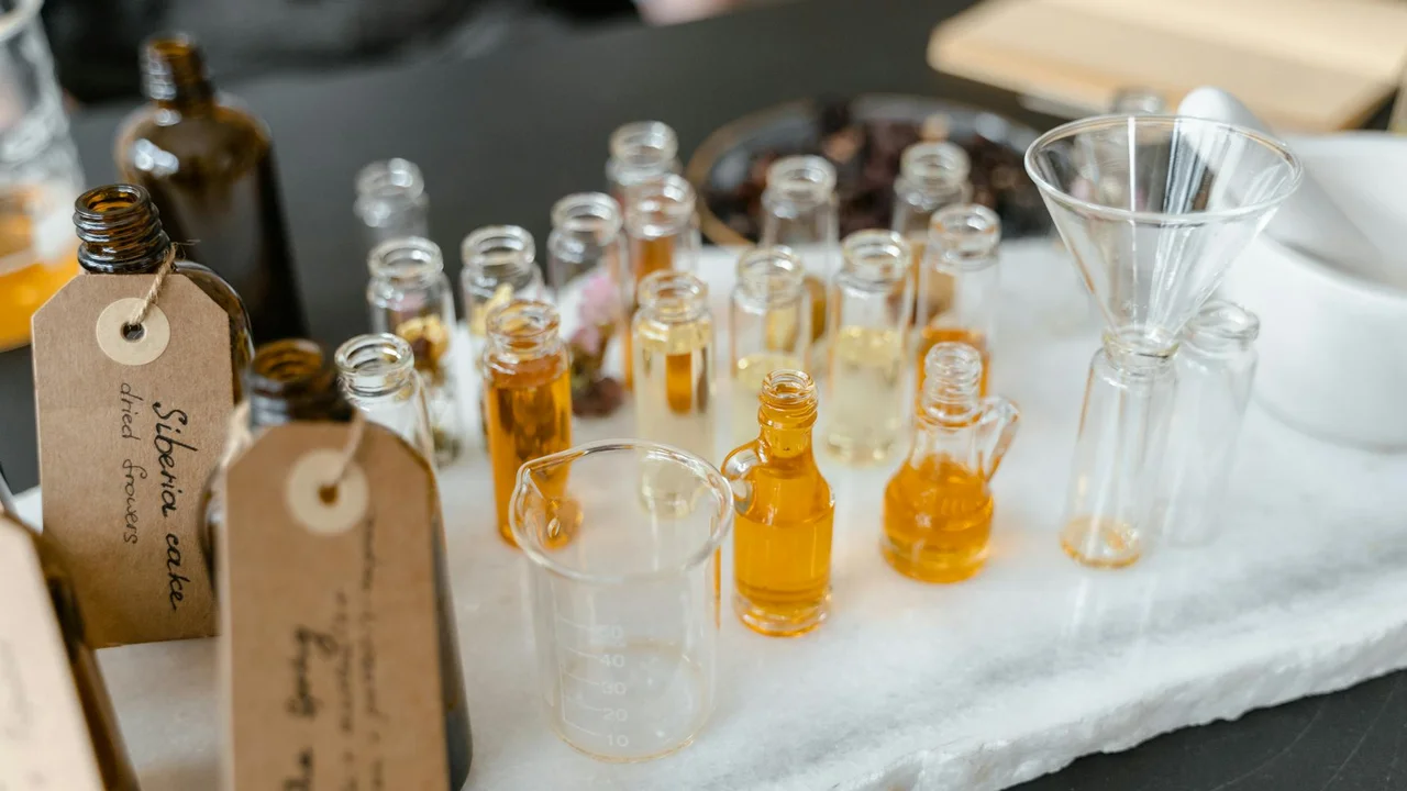 Assorted small amber glass bottles with droppers and a glass beaker arranged on a marble surface, suggesting a lip oil and gloss setup.