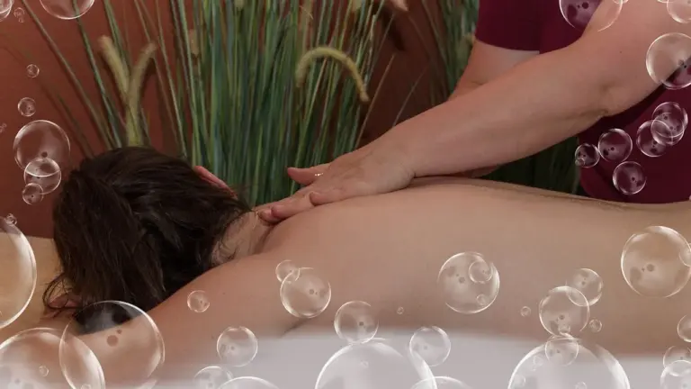 A person lies face down on a massage table while a therapist rubs their back; bubbles float around in a spa-like setting.