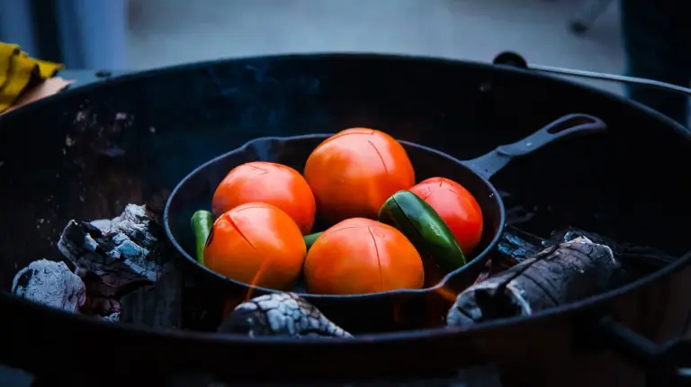 Cast iron cookware over a charcoal grill with a small pan containing red-orange tomatoes and green peppers, surrounded by ashes.