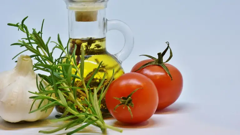 A bottle of olive oil surrounded by garlic, tomatoes, and rosemary on a white background
