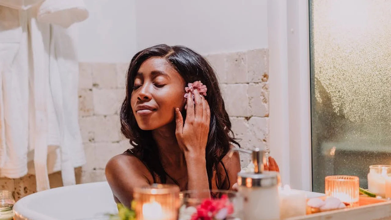 A woman in a bathroom applies skincare product to her face, surrounded by warm candles and a spa-like setting.