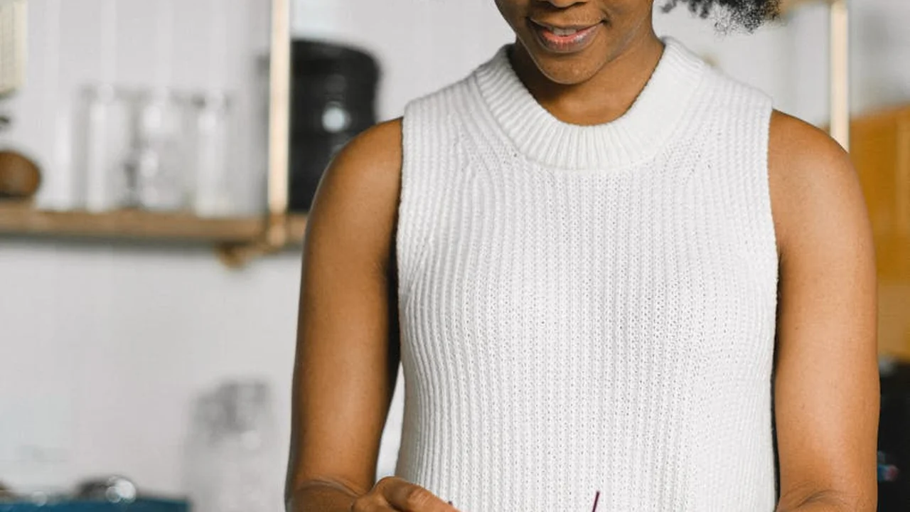 A person wearing a white sleeveless knit sweater stands in a bright kitchen with shelves in the background.