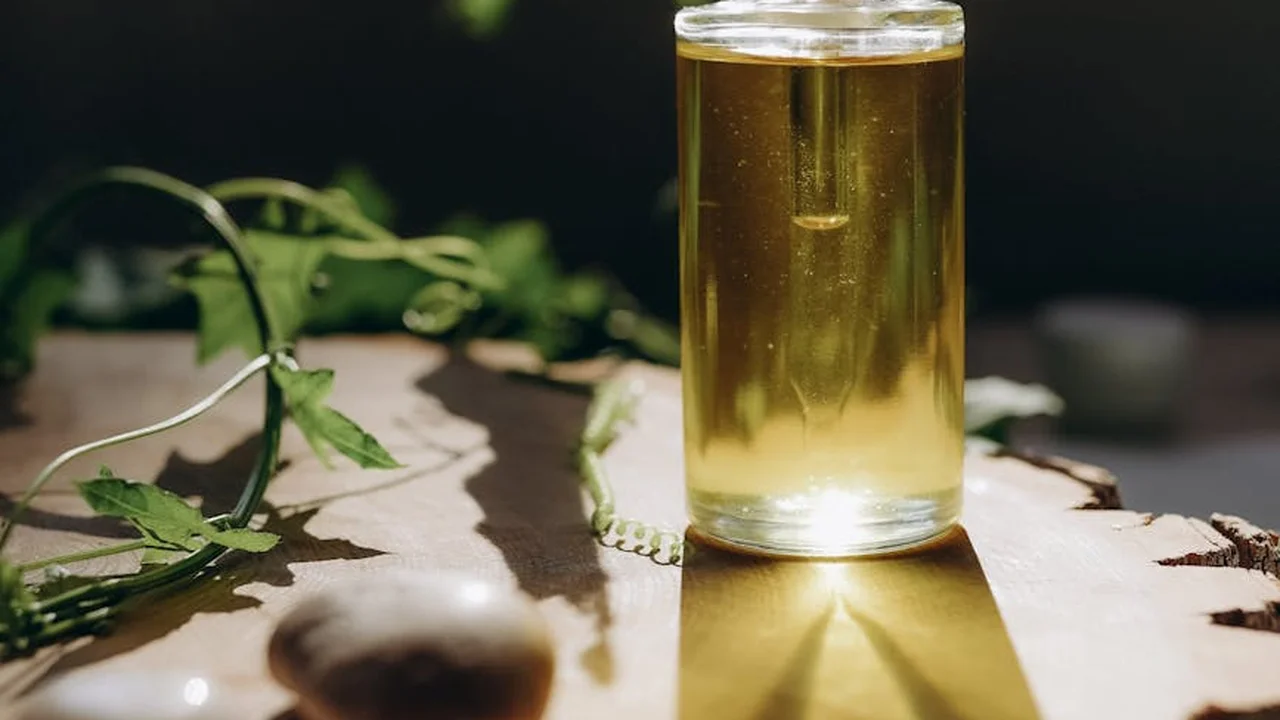 Clear glass bottle of light yellow oil on a wooden surface with green leaves in the background.