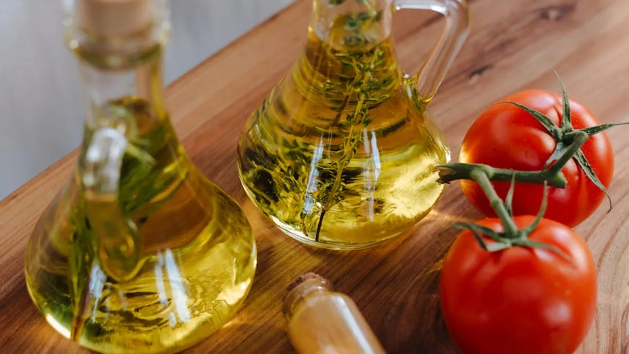 Two glass bottles of olive oil with sprigs inside, placed on a wooden surface beside ripe red tomatoes