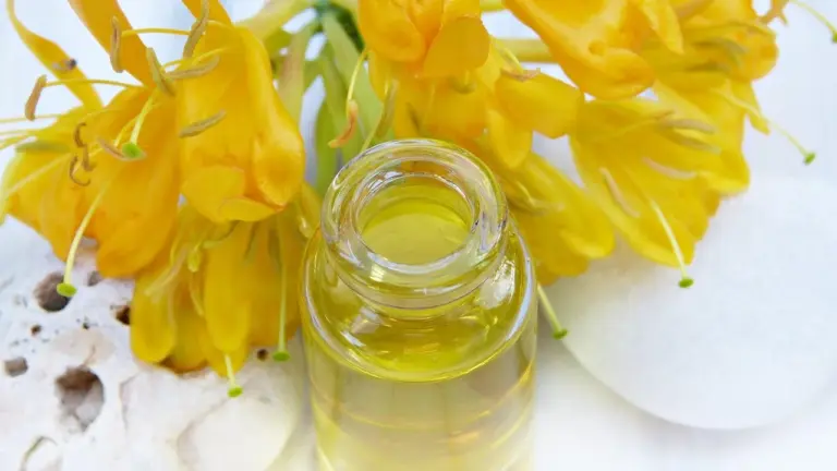 Close-up of a glass bottle of golden cleansing oil with bright yellow flowers in the background on a light surface.