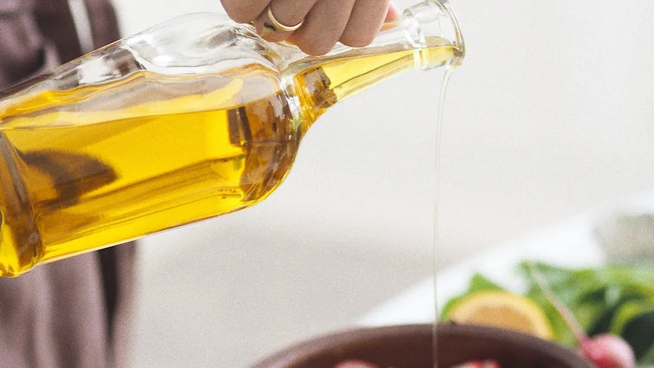 A hand pours golden oil from a glass cruet, with a fresh salad in the blurred background.