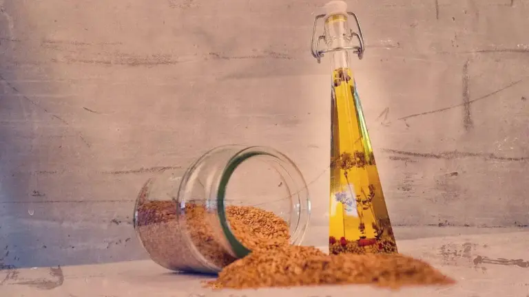 A clear glass bottle of golden oil stands upright beside an overturned glass jar with brown sugar spilled on a rustic wooden surface.
