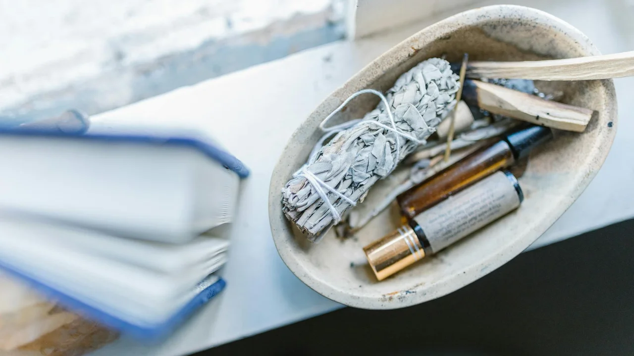 Ceramic dish containing essential oil bottles and a bundle of dried herbs on a desk beside an open book.