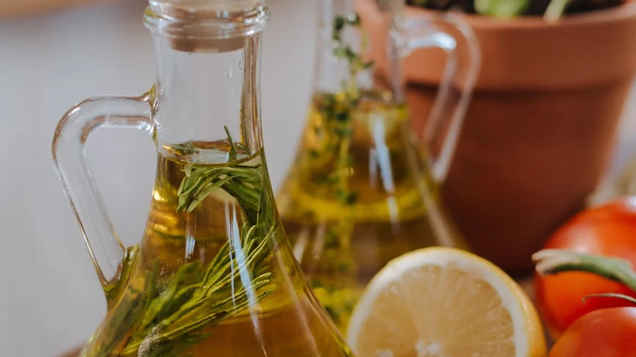 Close-up of glass oil bottles with fresh herbs, a lemon slice, and tomatoes in a kitchen setting.