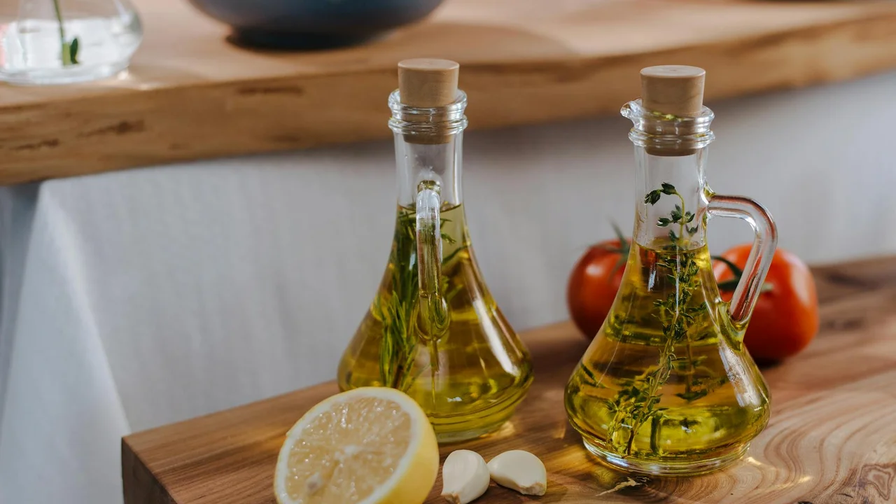 Two glass bottles of olive oil with herbs on a wooden kitchen board, accompanied by a lemon slice and garlic cloves