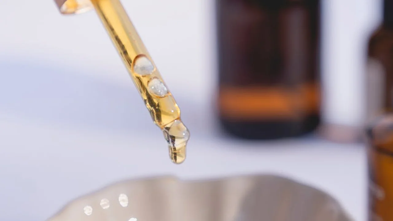 Close-up of a glass dropper releasing a droplet of golden botanical oil with blurred bottles in the background.