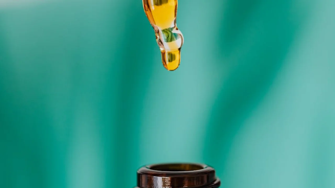 Close-up of an oil drop hanging from a dropper above a small vial, with a green, out-of-focus background.