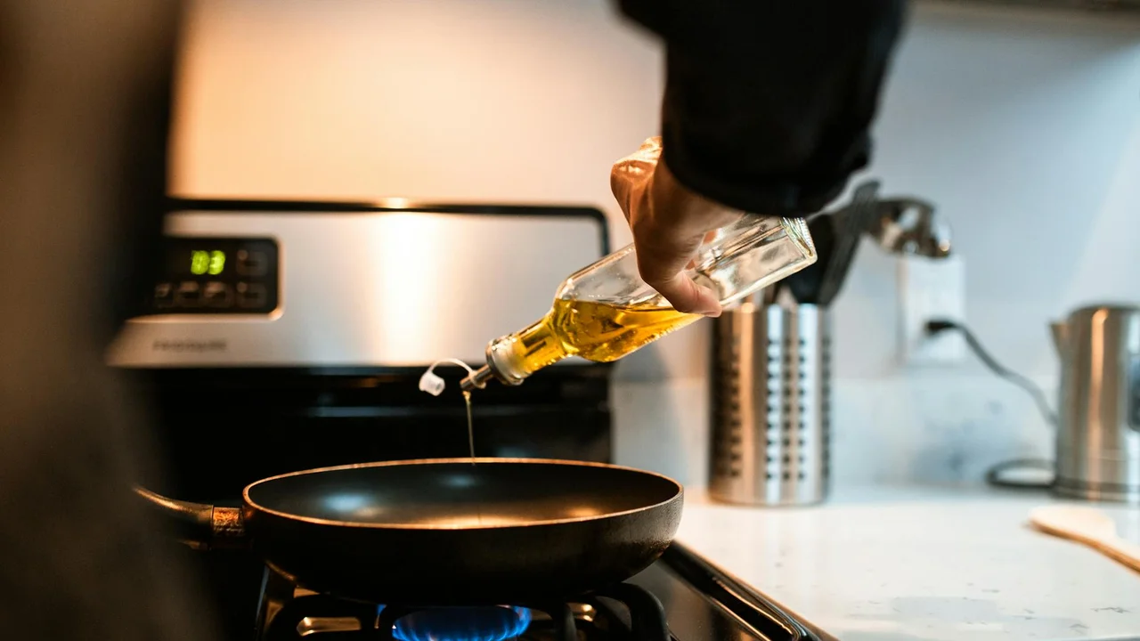 A hand holding a bottle of cooking oil and pouring into a frying pan on a gas stove in a kitchen.